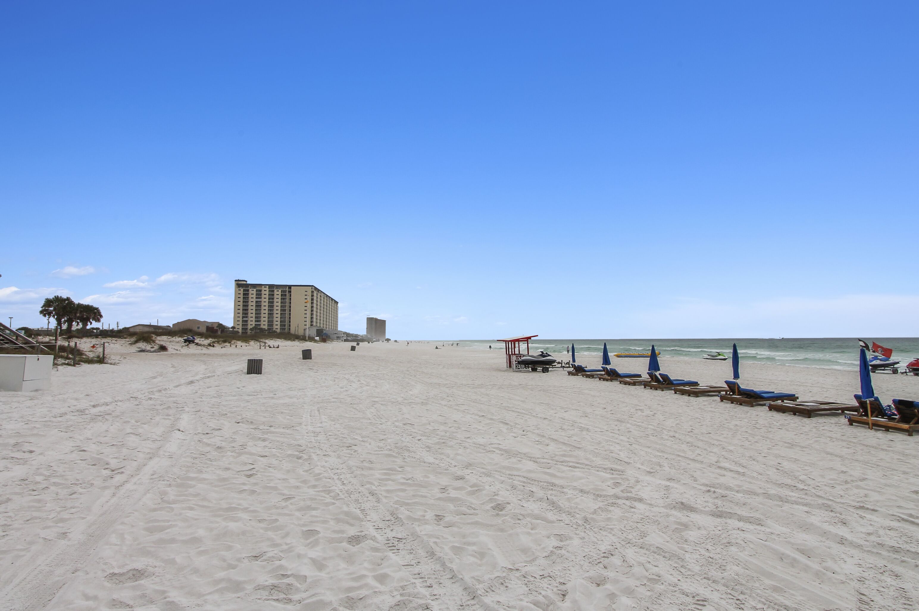 A beach with a row of blue beach chairs and umbrellas, a building in the background, and a clear blue sky.