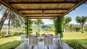 Een overdekt terras met een eettafel voor twee personen, omringd door witte stoelen, met uitzicht op een weelderig landschap met bomen en heuvels.