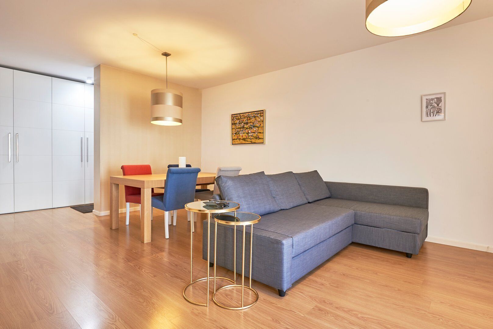 Ein modernes Wohnzimmer mit einem grauen Ecksofa, einem kleinen runden Couchtisch, einem Esstisch mit Stühlen und einer Pendelleuchte.