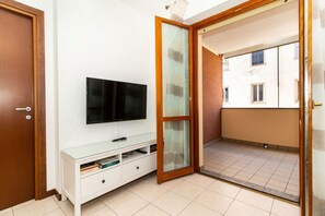 A room with a TV on a white stand, a sliding glass door leading to a balcony, and a wooden door.