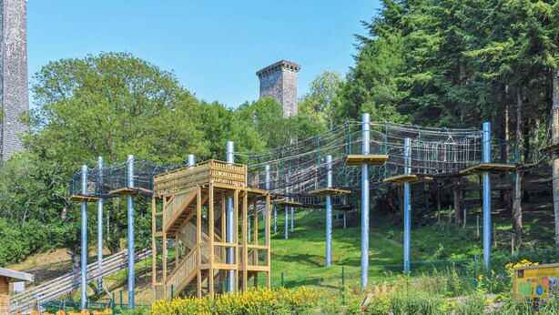 Un parque infantil con una estructura de madera para escalar, un puente de cuerda y una torre de piedra al fondo.