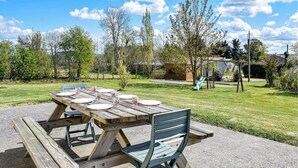 Una mesa de picnic con platos y vasos, sobre una superficie de concreto en un área verde con árboles y un parque infantil al fondo.