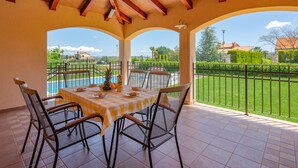 Eine überdachte Terrasse mit Esstisch, mit Blick auf einen Swimmingpool und einen umzäunten Garten.