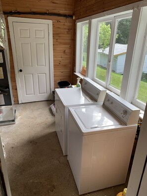 A compact laundry room with a washer and dryer, wooden walls, a white door, and windows overlooking a yard. - Completely Remodeled Country Cottage  (Knoxville)