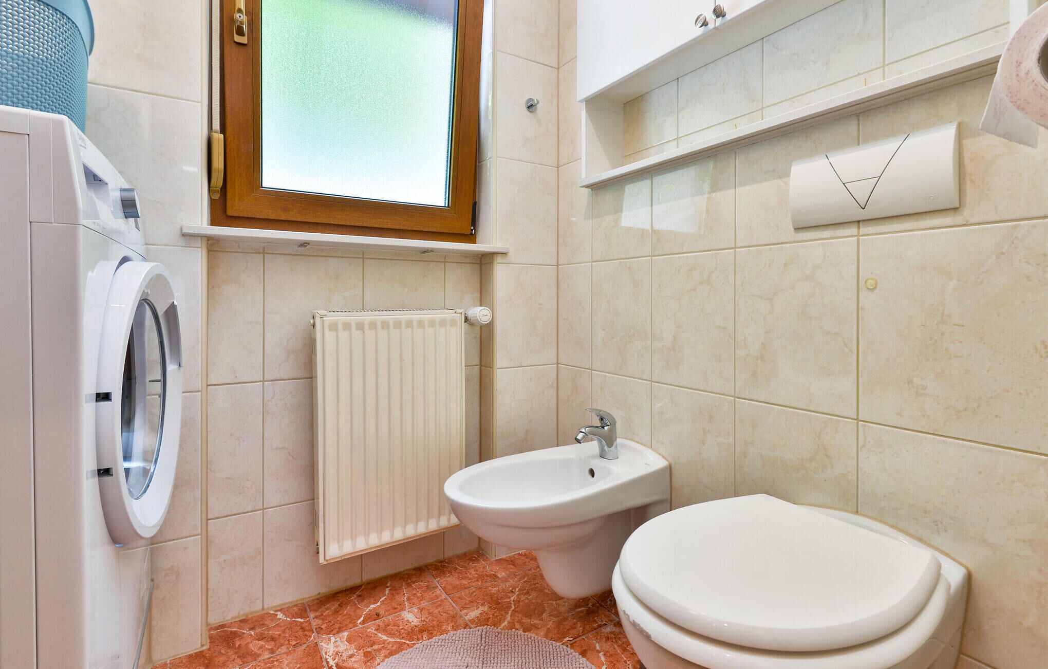 Une salle de bain équipée d’un lave-linge, d’un bidet, de toilettes, d’un radiateur et d’une fenêtre à vitre dépolie.