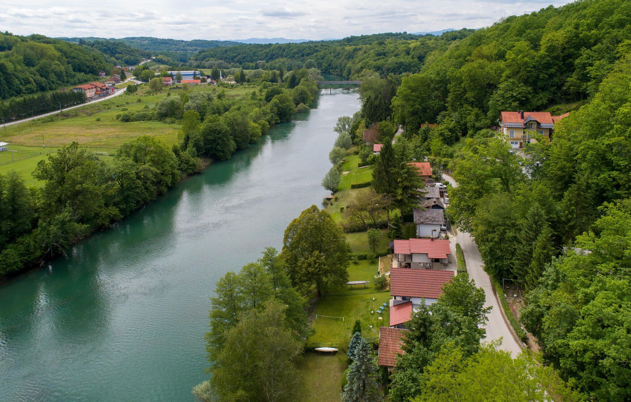 A riverside village with houses along the riverbank, a bridge in the distance, and a hilly landscape.