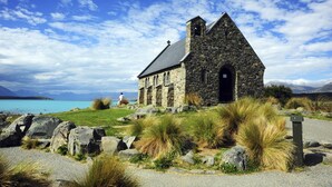 A stone church with a stone roof, situated by a body of water with mountains in the background. - Matariki Star - 1 Bedroom Suite A (Lake Tekapo)