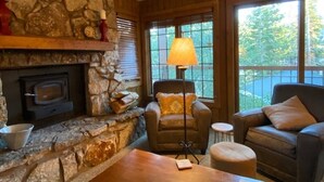 Una sala de estar acogedora con chimenea de piedra, un sofá de cuero cómodo, una mesa de centro de madera y un ventanal con vistas a árboles.
