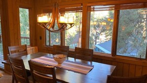 Un comedor con una mesa y sillas de madera, una araña y ventanas amplias con vistas a árboles y edificios.