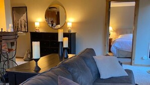 Habitación de hotel moderna con un sofá gris, un espejo redondo, un escritorio con una silla y una cama al fondo.