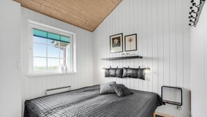 Een slaapkamer met een bed, een raam met zonwering, een plank met decoratieve objecten en een wandlamp.