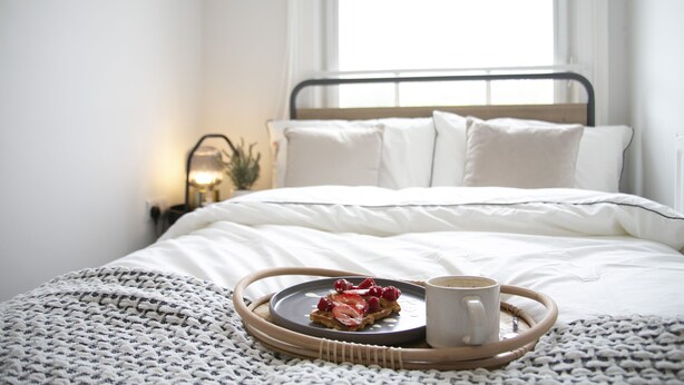A neatly made bed with white linens, a tray with waffles and berries, and a cup of coffee on a woven tray.