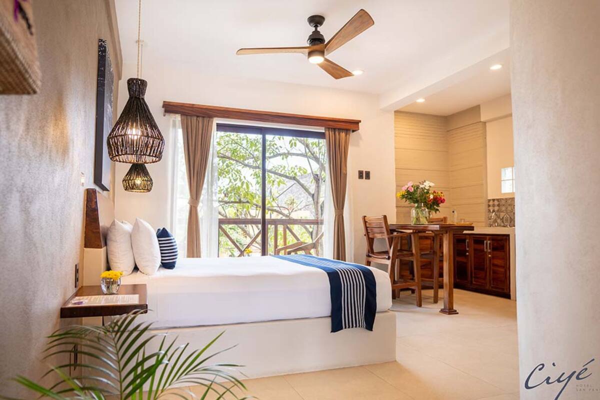A hotel room with a bed, a ceiling fan, a dining area with a table and chairs, and a view of the outdoors.
