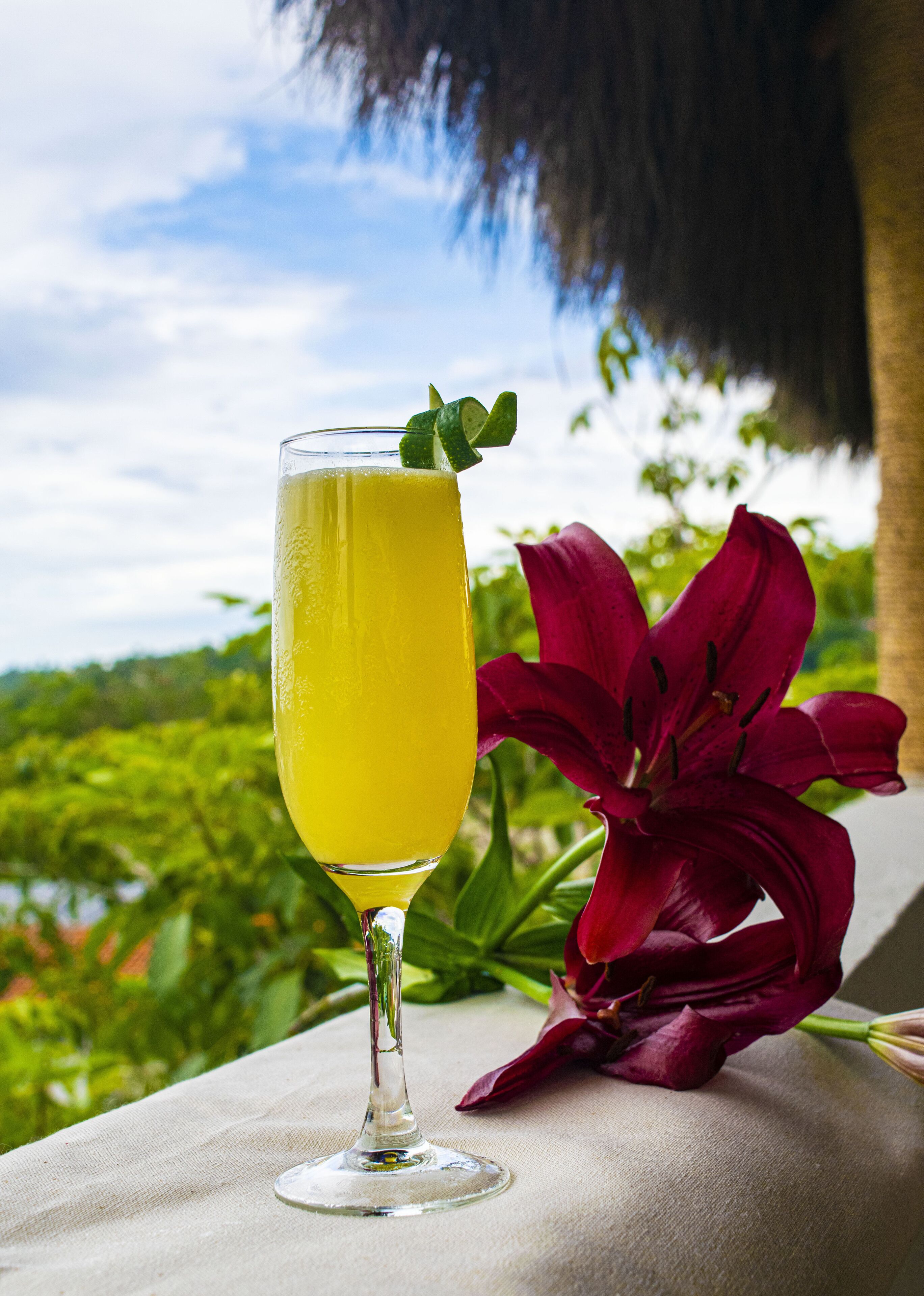 A clear glass of yellow beverage with a lime garnish on a table with a red flower beside it.