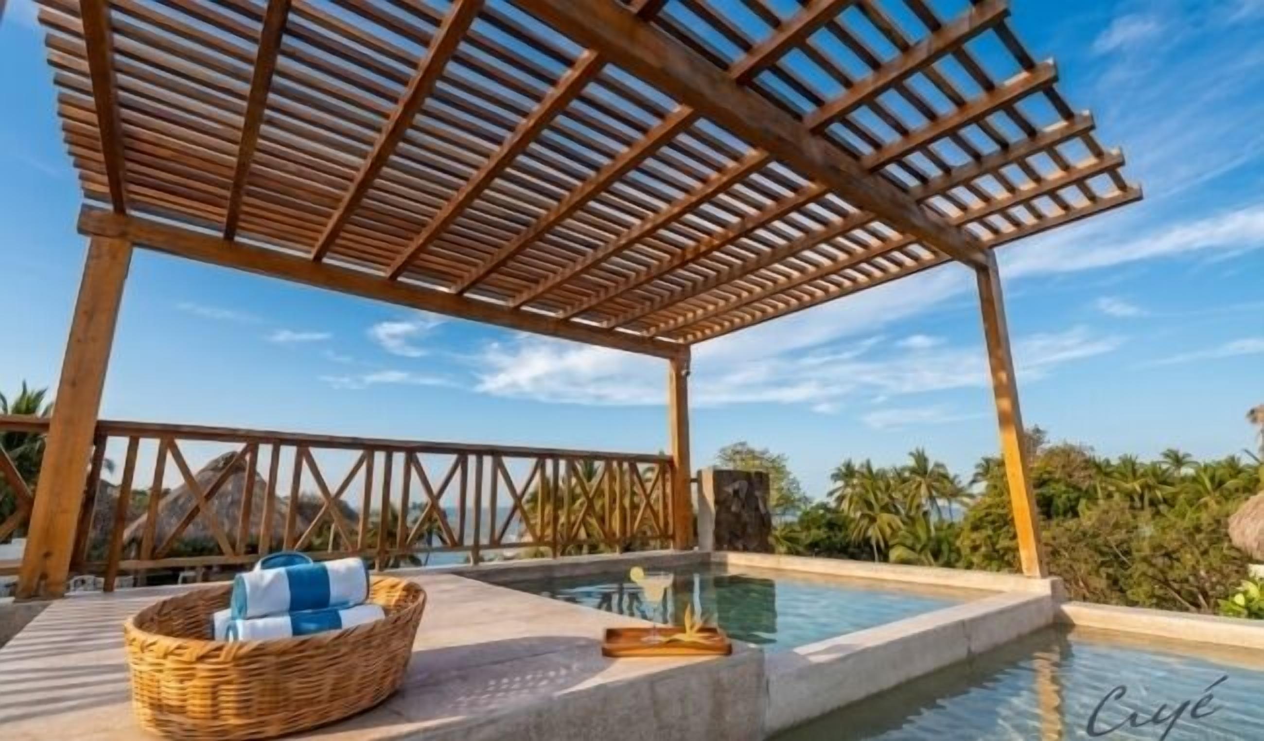 An outdoor pool area with a wooden pergola, a wicker basket with towels, and a clear view of the surrounding landscape.