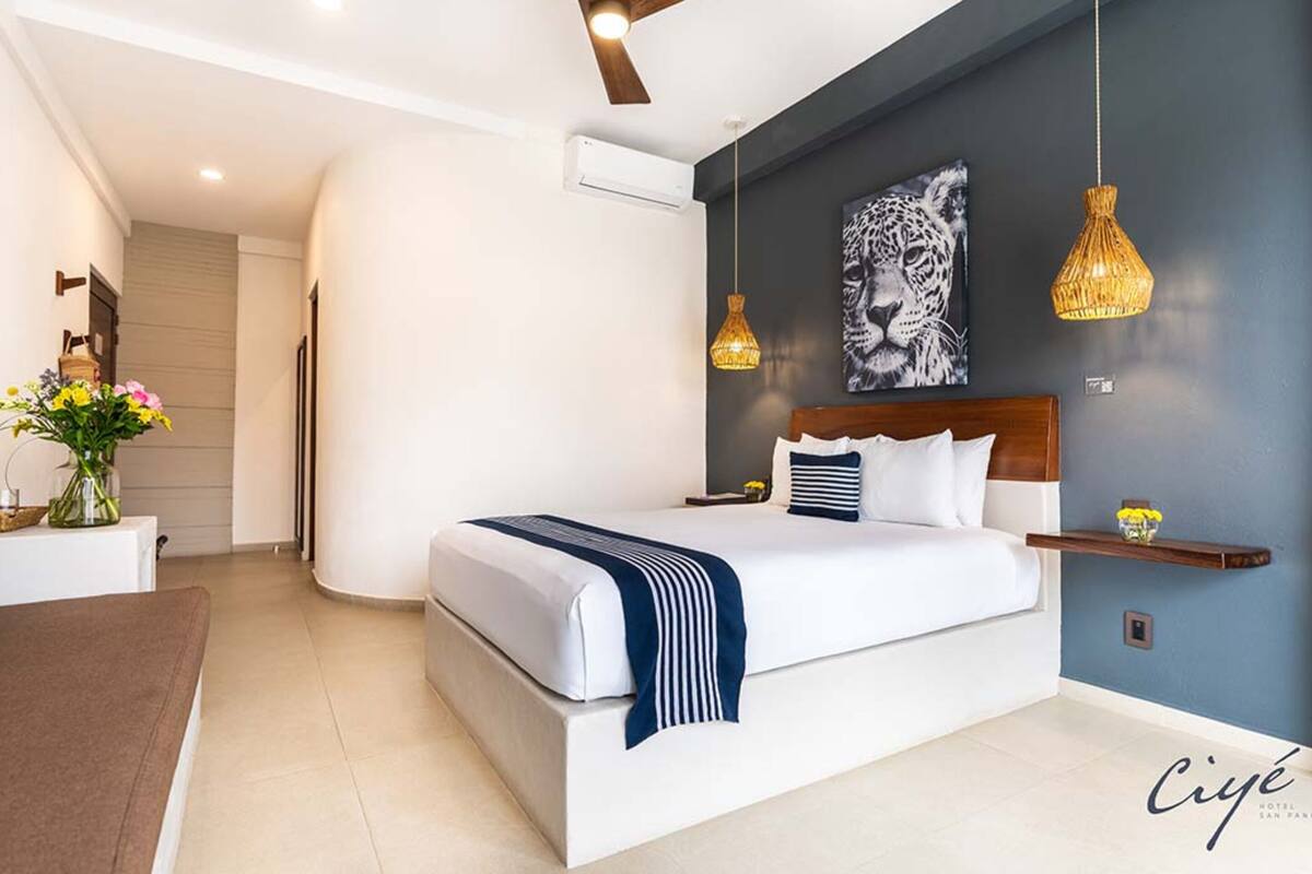 A modern hotel room with a large bed, a ceiling fan, a wall-mounted air conditioner, and a decorative painting.