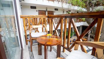 Un área de estar al aire libre con mesasillas y sillas de madera, una barandilla de vidrio y vistas a un edificio con balcones y plantas.