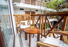 Un área de estar al aire libre con mesasillas y sillas de madera, una barandilla de vidrio y vistas a un edificio con balcones y plantas.