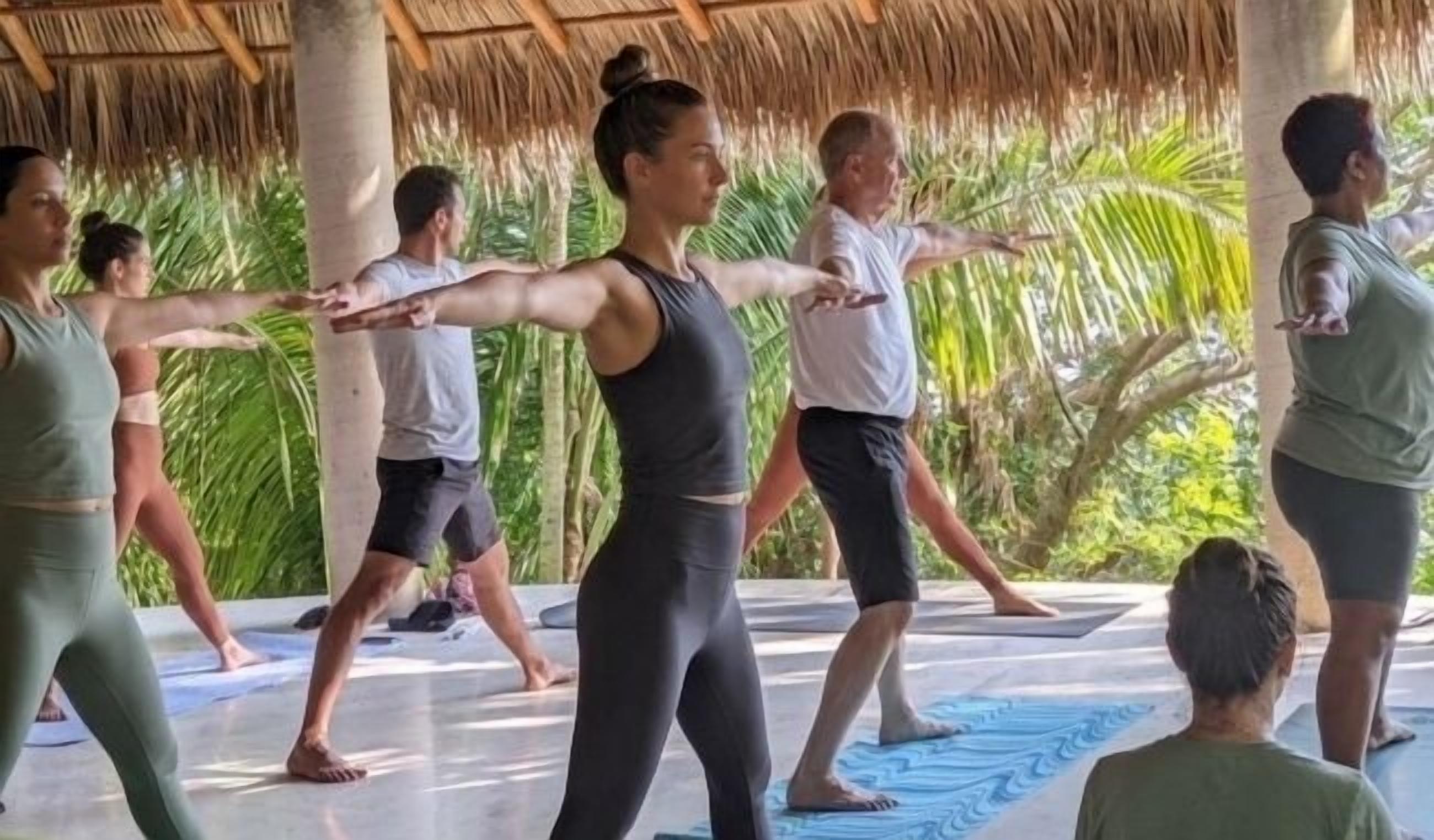 Un grupo de personas realizando ejercicios de yoga al aire libre, con una estructura de techo de paja y palmeras al fondo.