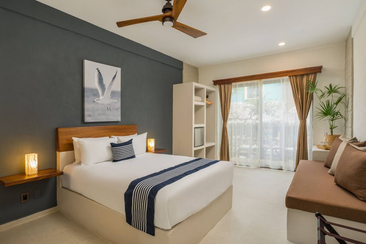 A modern hotel room with a large bed, a ceiling fan, a bedside table with a lamp, a bookshelf, and a seating area with a sofa and a coffee table.