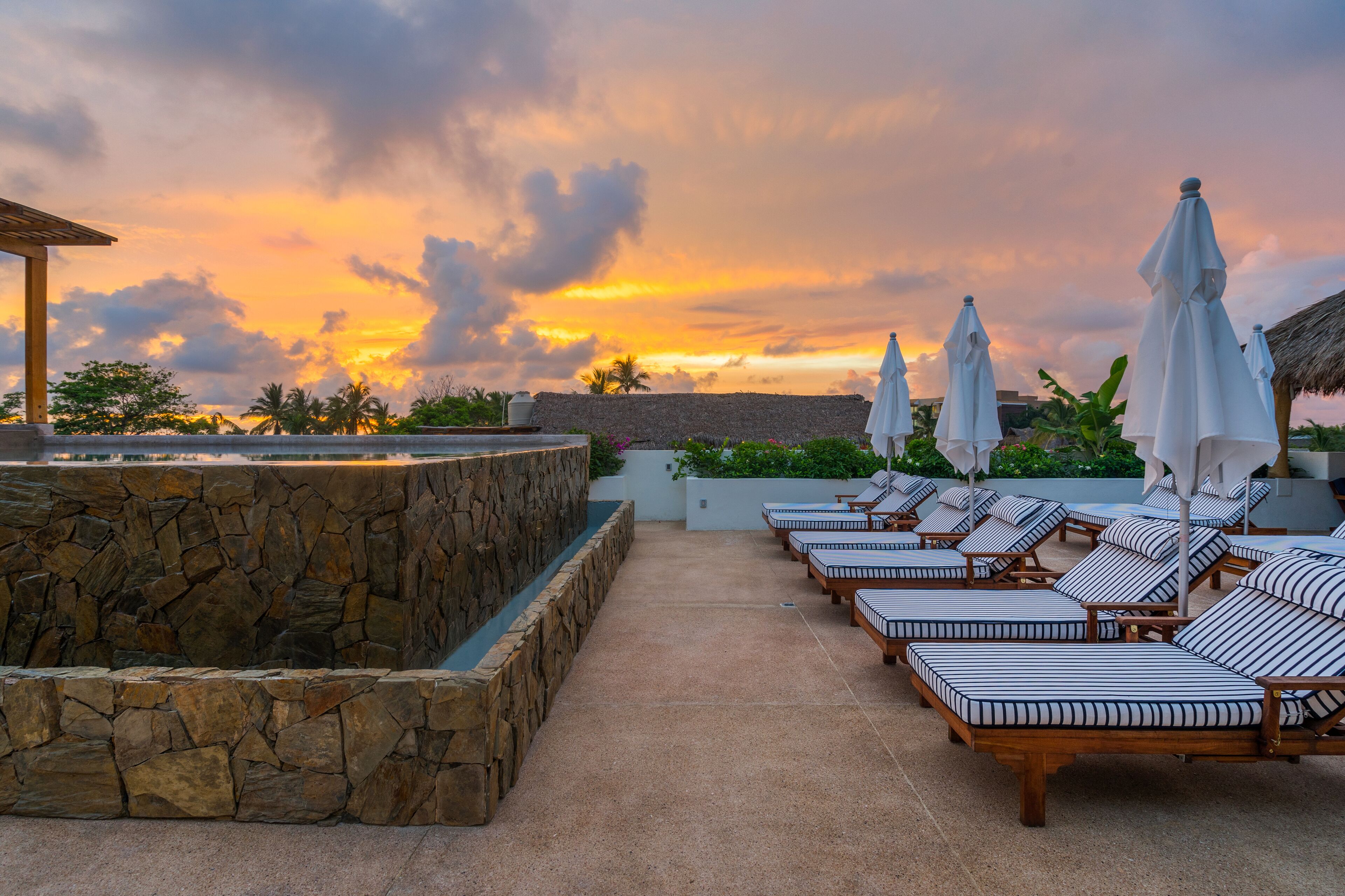 Un área de lounge en la azotea con paredes de piedra y sillones con cojines a rayas, bajo un cielo al atardecer.