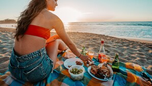Una persona sentada sobre una manta de playa con comida y bebidas.