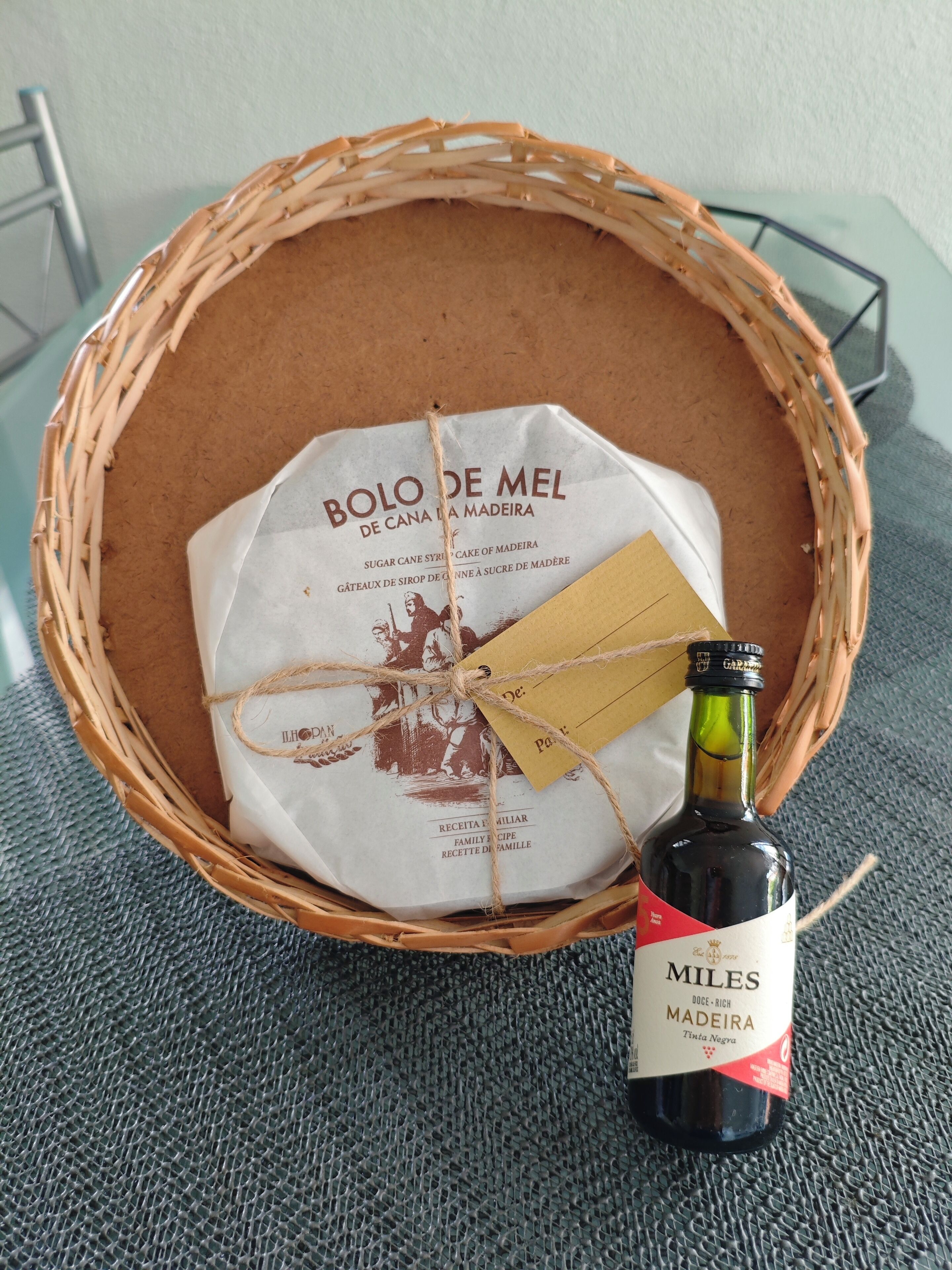Un panier en osier contenant une bouteille de vin de Madère de la marque Miles, une étiquette et un morceau de papier avec du texte.