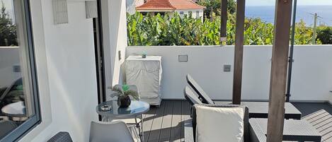 Un espacio al aire libre cubierto con sofás y sillones, una pequeña mesa con una planta, y vistas al mar y un edificio de techo rojo al fondo.