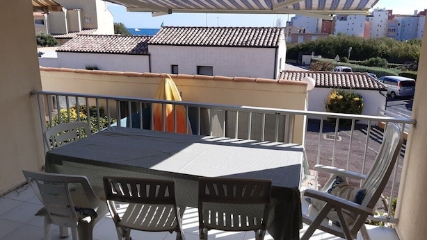 Ein Balkon mit Tisch und Stühlen, einem gestreiften Sonnenschirm und Blick auf Gebäude und Meer.