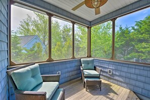 Un porche cubierto con mobiliario de mimbre y ventanas amplias que ofrecen vistas a una casa y árboles.