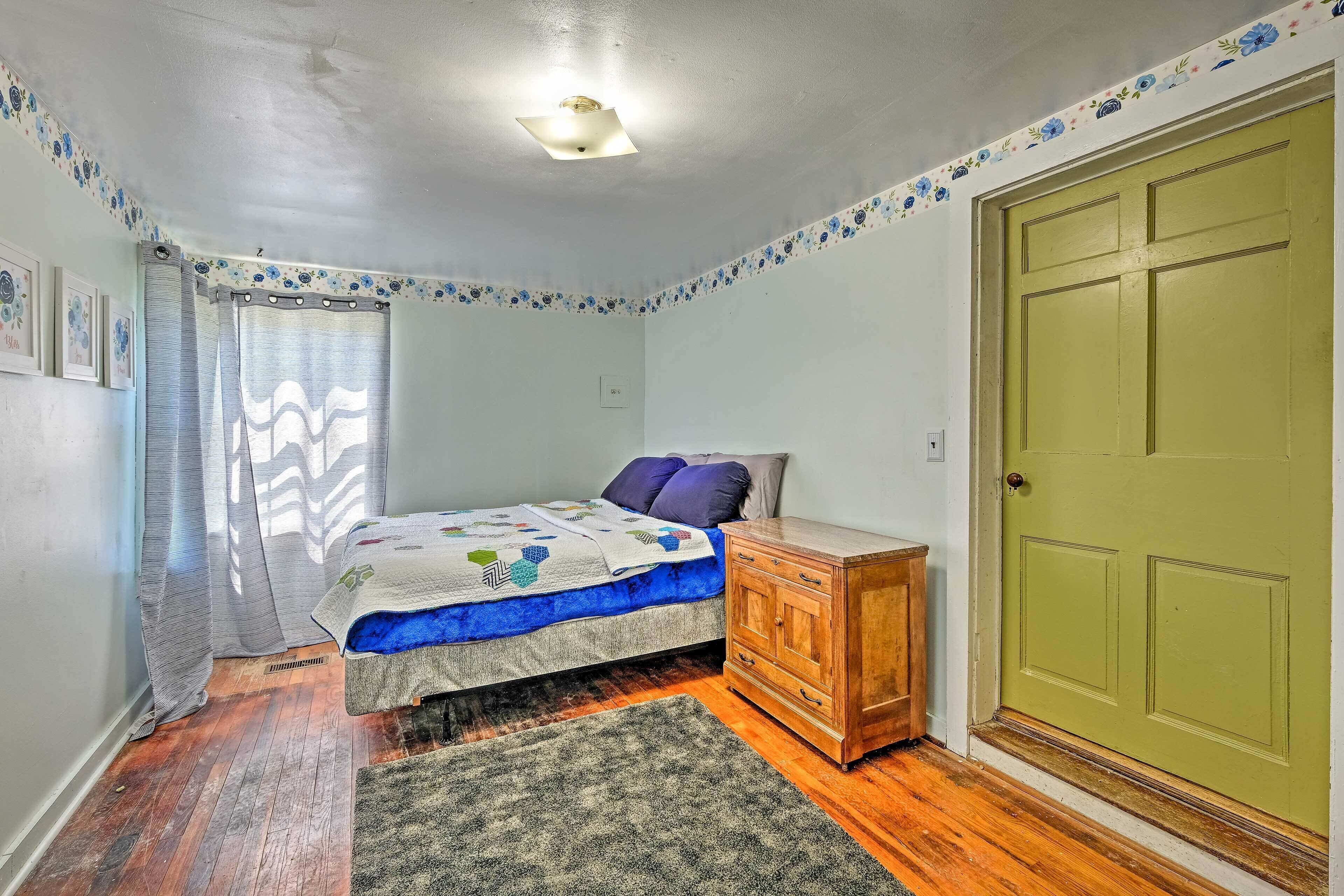 A bedroom with a bed, a wooden dresser, a green door, and a window with curtains.