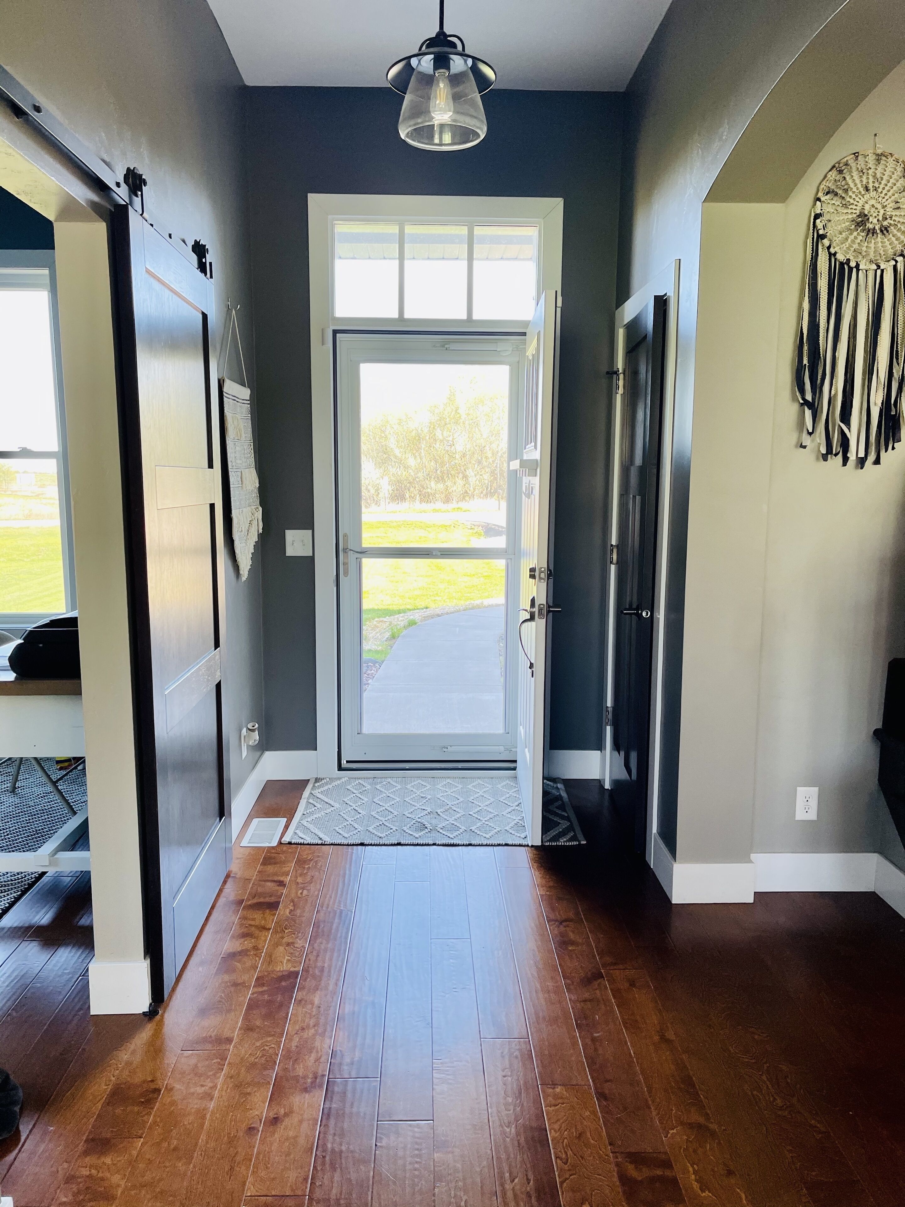 A hallway with a wooden floor, a sliding door leading to a bright outdoor area, and a hanging light fixture.