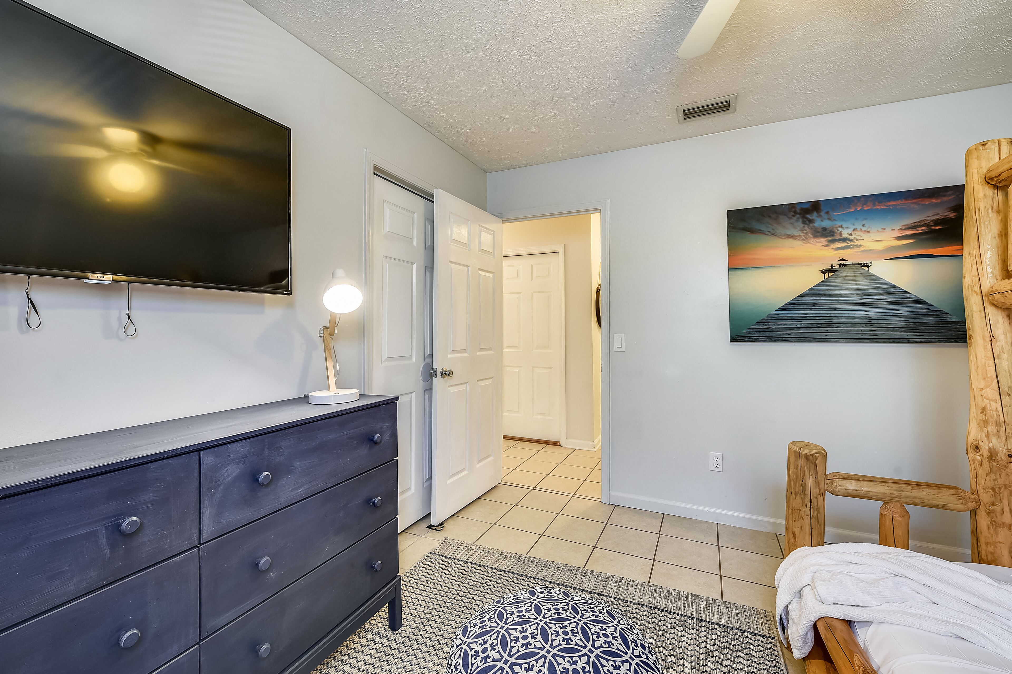 A bedroom with a large flat-screen TV, a wooden chair, a patterned rug, and a painting of a sunset over water.
