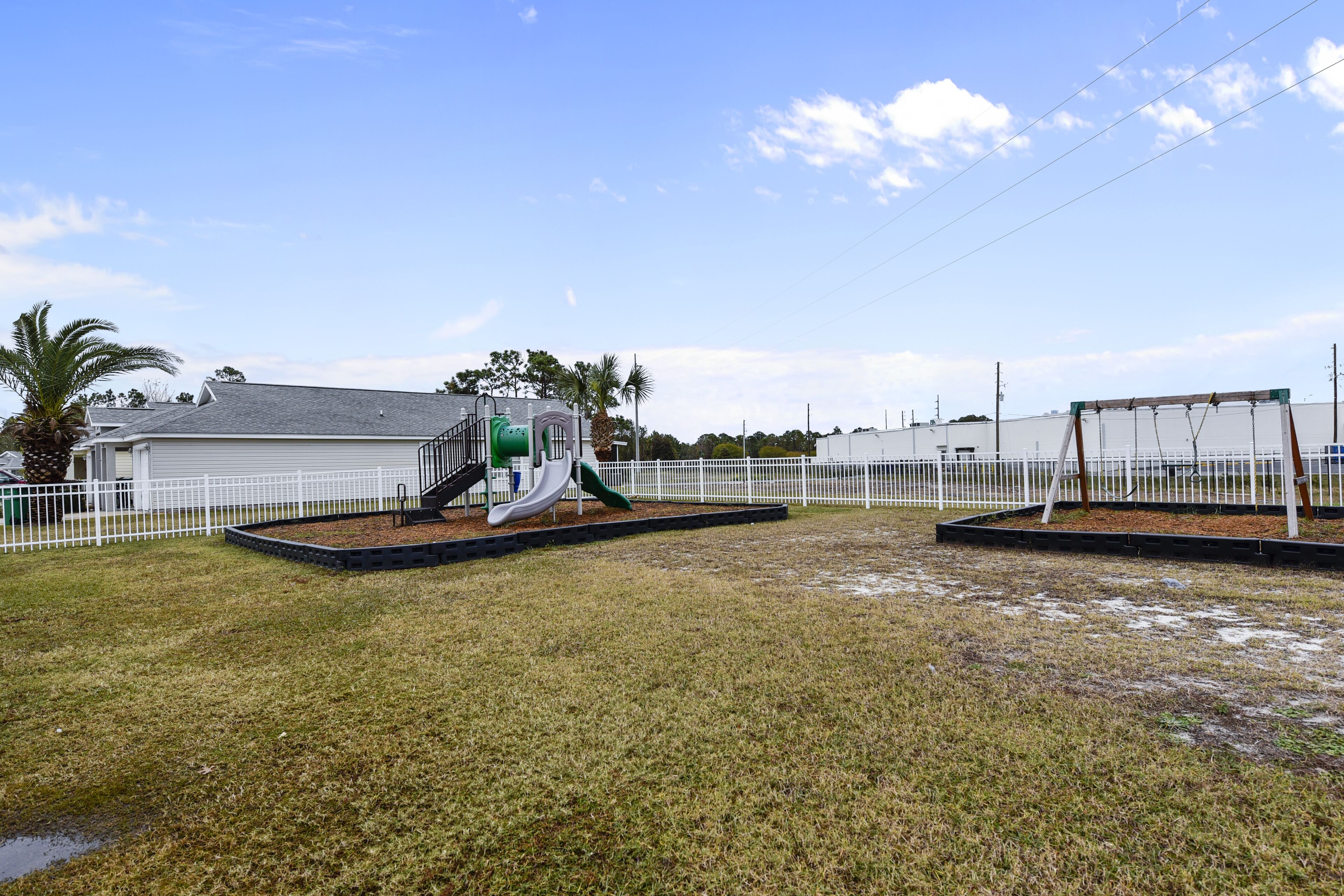 A playground with a slide and swings, surrounded by a white fence and palm trees.