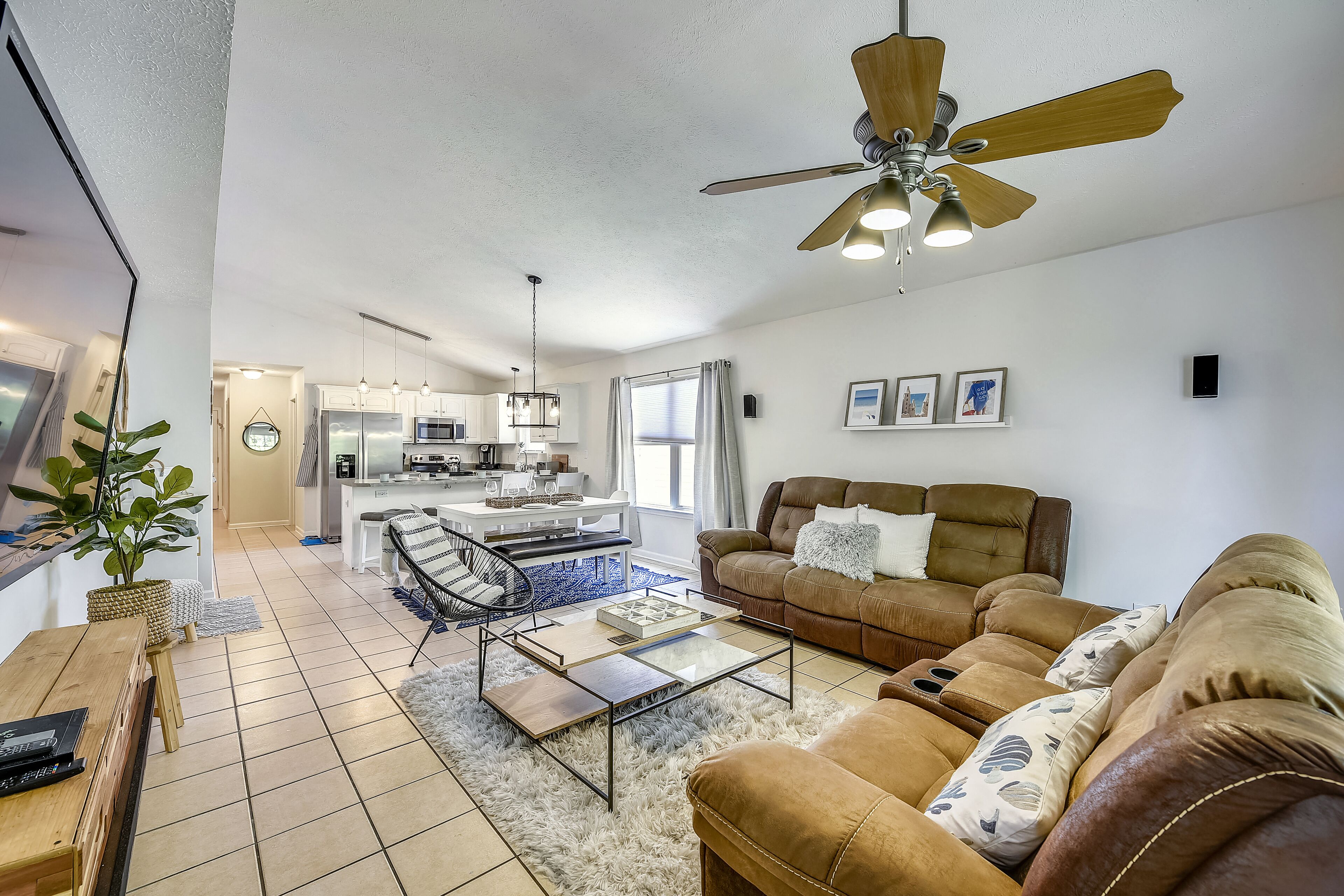 A modern living room with a sofa, coffee table, and a ceiling fan. There is a kitchen in the background with white cabinets and a dining area.