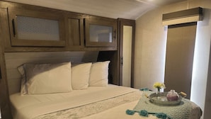 Uma cama bem arrumada com colcha estampada, uma mesa de cabeceira com um vaso e um frasco, e uma janela com cortinas.