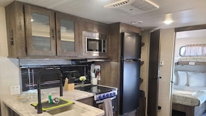Uma cozinha compacta com micro-ondas, fogão e frigorífico, uma pia pequena e uma janela com cortinas estampadas.