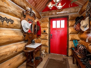 A log cabin interior with a red door, wooden walls, a chandelier, and various hats and boots displayed.