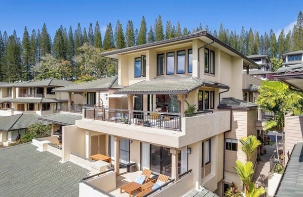 A multi-story residential building with a balcony, large windows, and a view of a forested area.