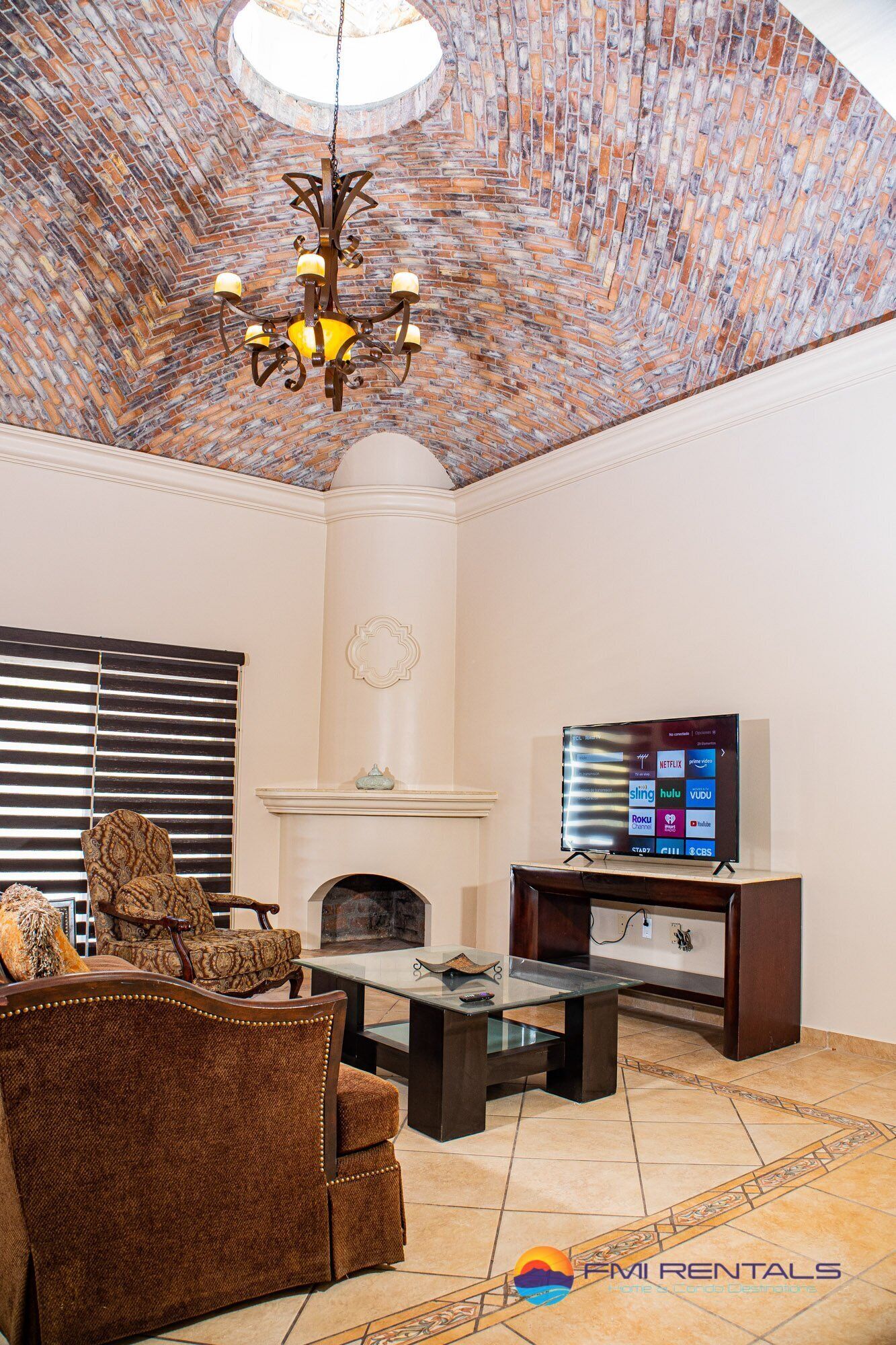 Een woonkamer met een bakstenen plafond, een open haard, een televisietoestel en een zithoek met een bank en fauteuils.