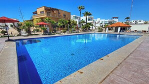A swimming pool with lounge chairs and umbrellas, surrounded by palm trees and modern buildings. - Casa Costa Diamante F27 (Puerto Peñasco)