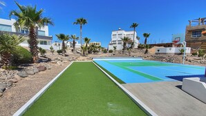 A courtyard with a green artificial turf and a blue sports court, surrounded by palm trees and modern buildings. - Casa Costa Diamante F27 (Puerto Peñasco)