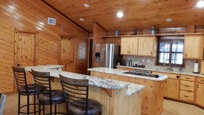 A wooden cabin interior with a kitchen area featuring a central island with bar stools, wooden cabinets, and a stainless steel refrigerator. - Hotel luxury in a Log Cabin (Rockport)