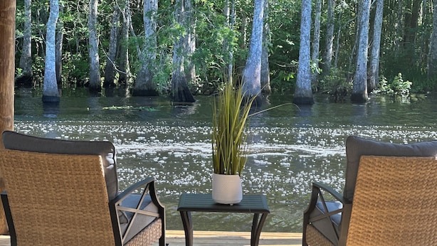 Een houten terras met twee rieten stoelen en een kleine tafel met een bloempot, met uitzicht op een kalm wateroppervlak en bomen op de achtergrond.