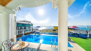 A pool area with a view of the sea, a covered seating area, and a table with a drink.