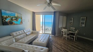 A living room with a beige sofa, a dining table with chairs, a ceiling fan, and a large window with a view of the beach and ocean.