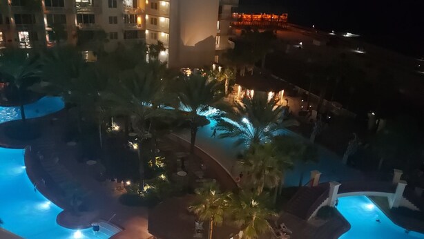 A nighttime view of a hotel pool area with multiple pools, palm trees, and a building in the background.