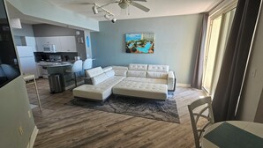 A modern living room with a sectional sofa, a dining area with a table and chairs, a kitchen with appliances, and a large window with a view of a pool and buildings.