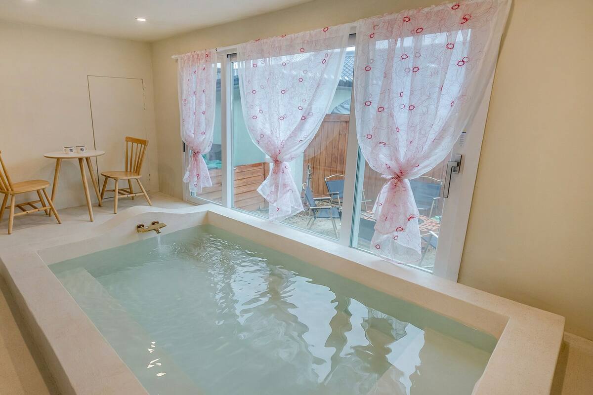 A room with a bathtub, two wooden chairs, a small table, and a window with curtains.