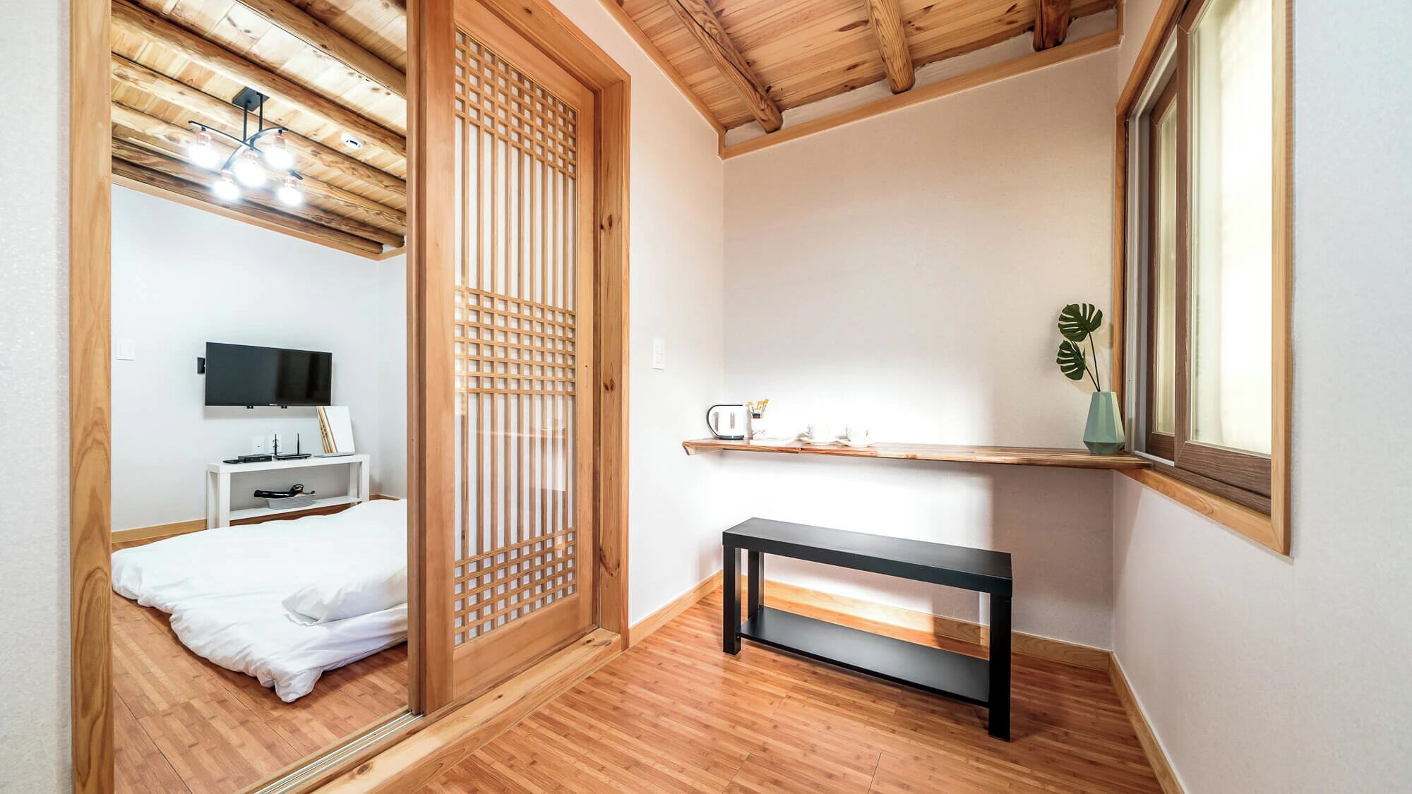 A room with a bed, a TV, a wooden partition, a small table, and a window with a plant.